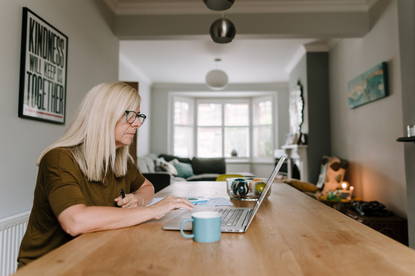 Image of a woman sitting at a table in front of a laptop, with a mug alongside.