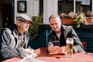 Two older adults looking at a mobile telephone. A cup and saucer, pint of beer, and newspaper are on the table in an outdoor setting.