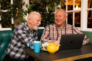 Two older males laughing, drinking tea, using a laptop.