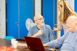 Two older adults sitting in front of a laptop. A younger adult is standing alongside explaining something.